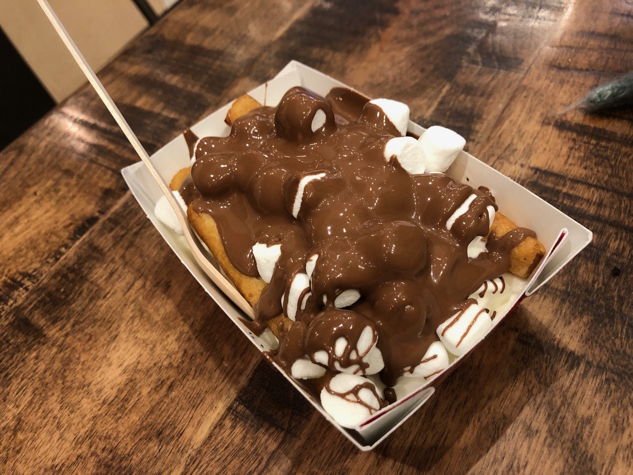 Dessert poutine in a cardboard container on a wooden table. Visible is a lot of chocolate sauce covering mini marshmallows and small churros-like fries